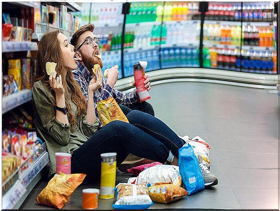 Couple sitting on the floor of a supermarket eating all kinds of snacks.